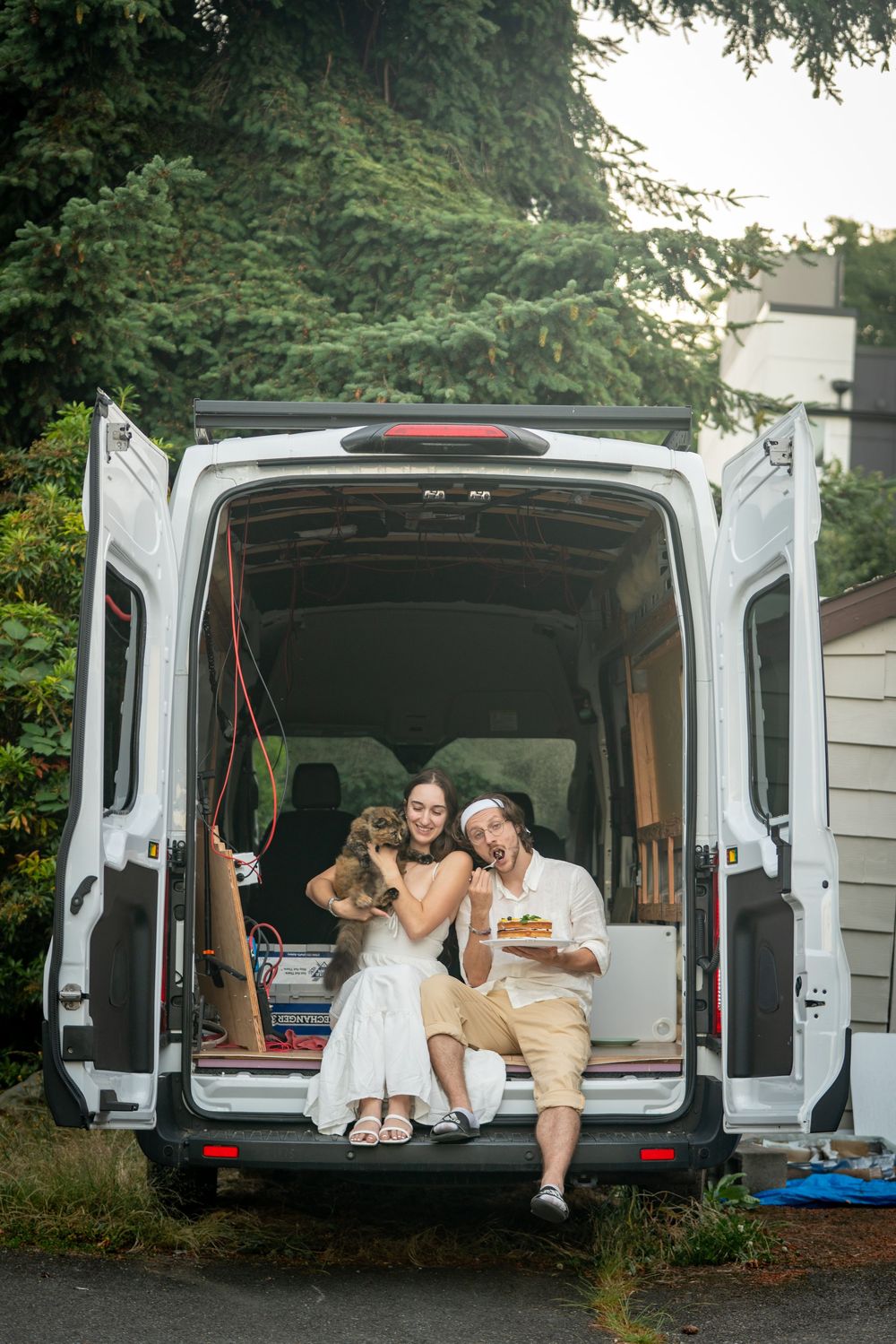 Happy newly weds sit in the open back of a white van with a cat, enjoying wedding cake outdoors.