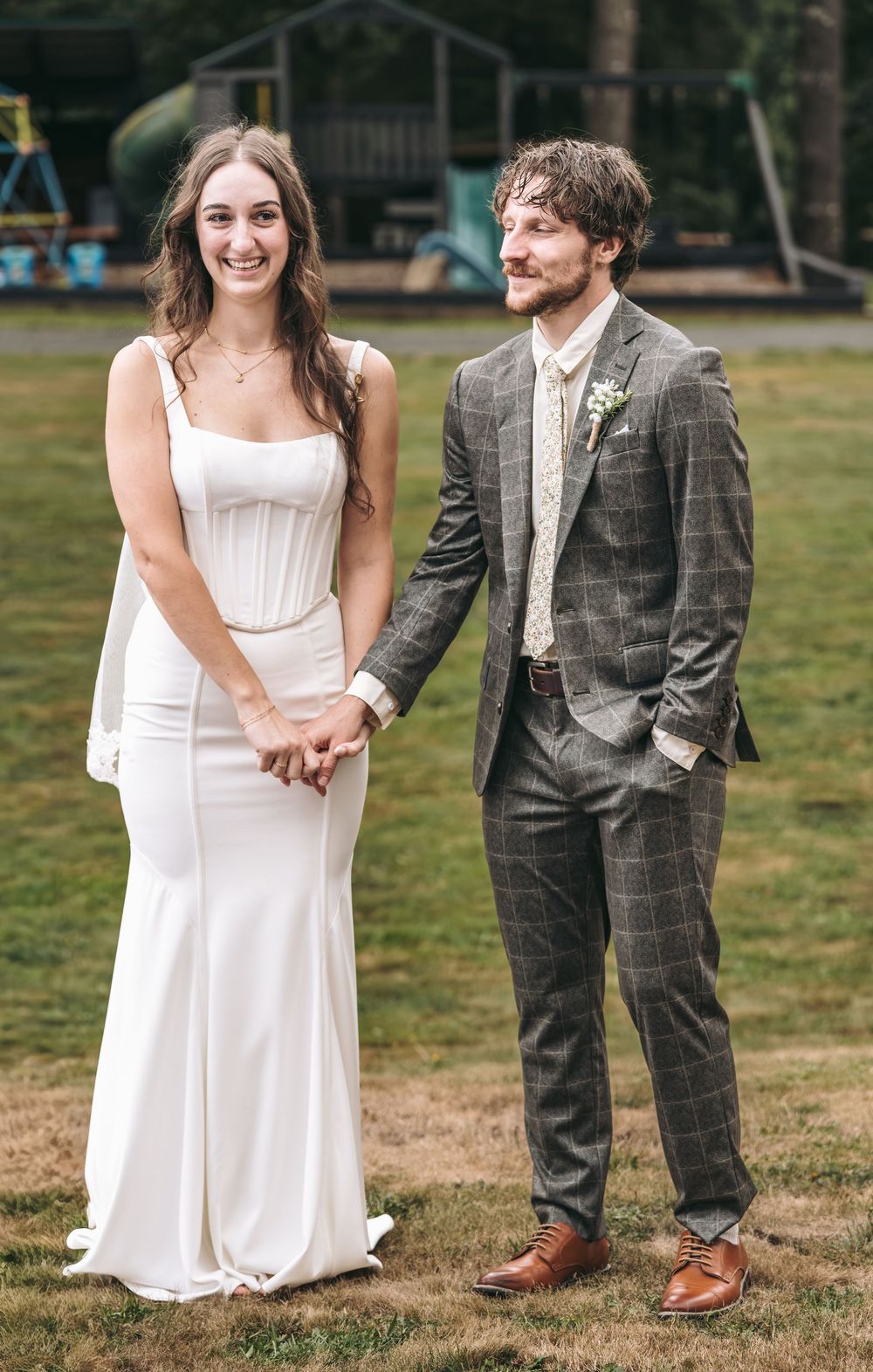 A smiling bride in a white gown holds hands with a groom in a gray plaid suit outdoors.