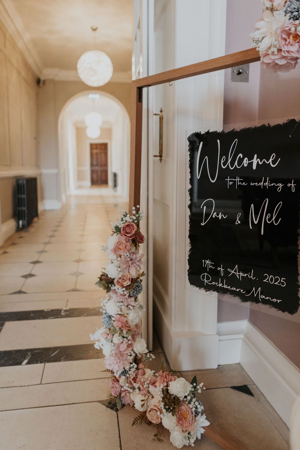 Welcome sign for Dan & Mel's wedding at Rockbeare Manor, April 2025, adorned with pink and white floral arrangement.