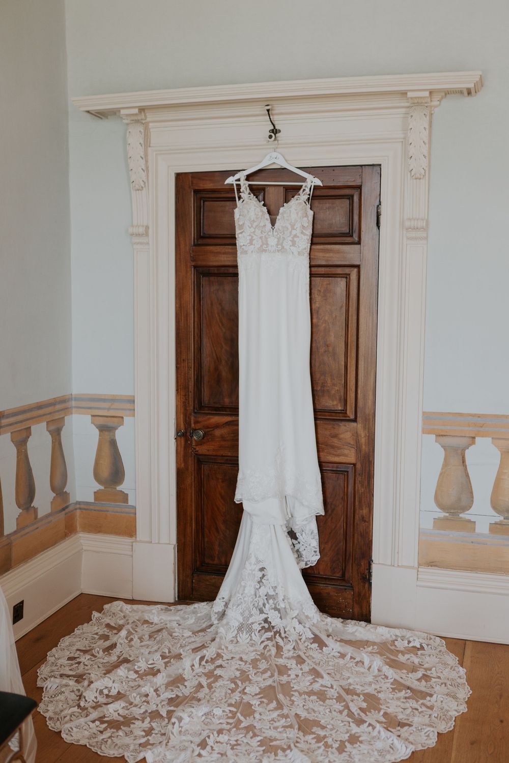 Elegant white lace wedding dress with long train hanging on a wooden door at Rockbeare Manor.