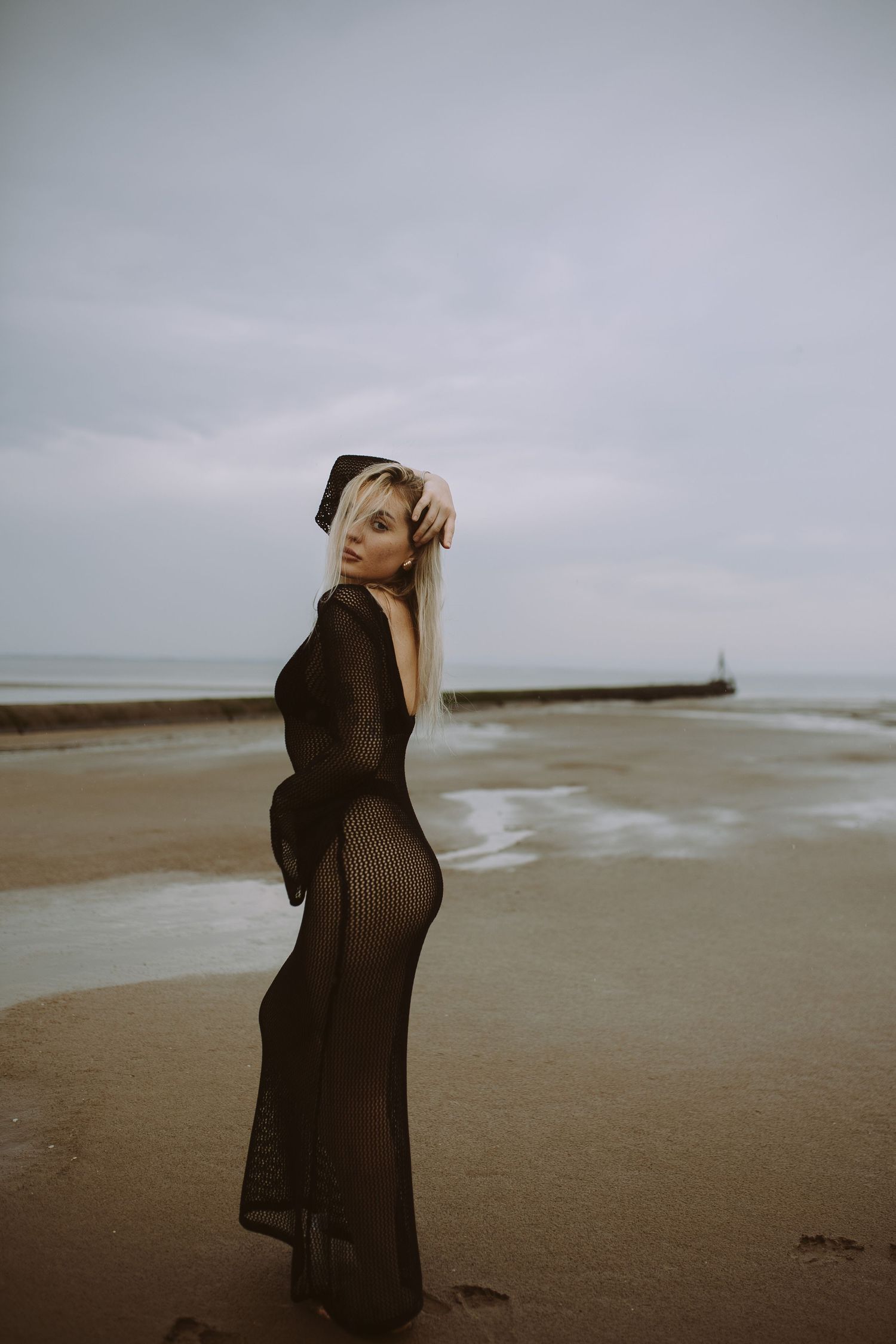 Silhouetted figure in dark clothing stands on a moody beach under overcast skies.