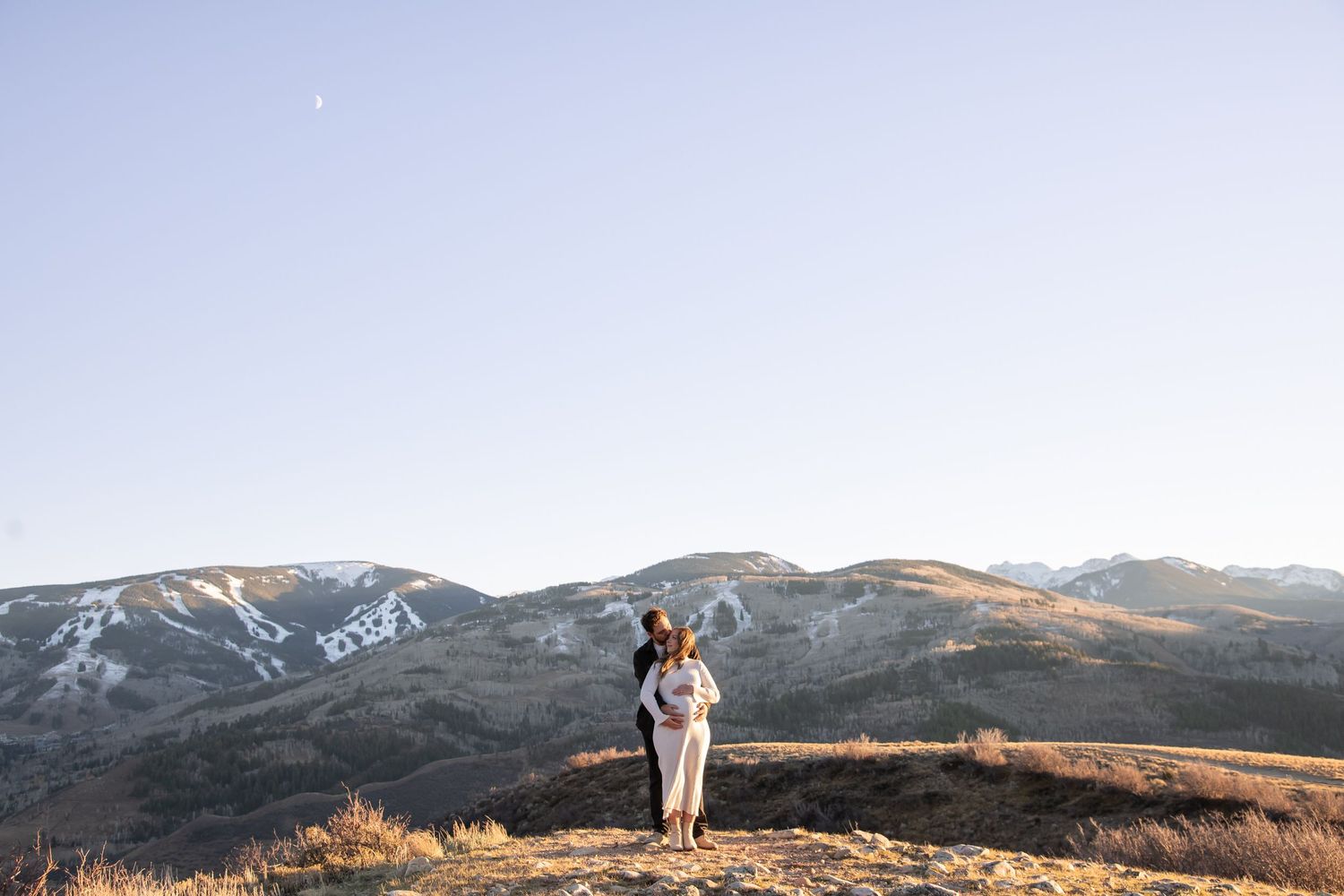 A couple stands atop a mountain ridge at sunset with snow-capped peaks and golden light in the background for a Vail maternity photo session