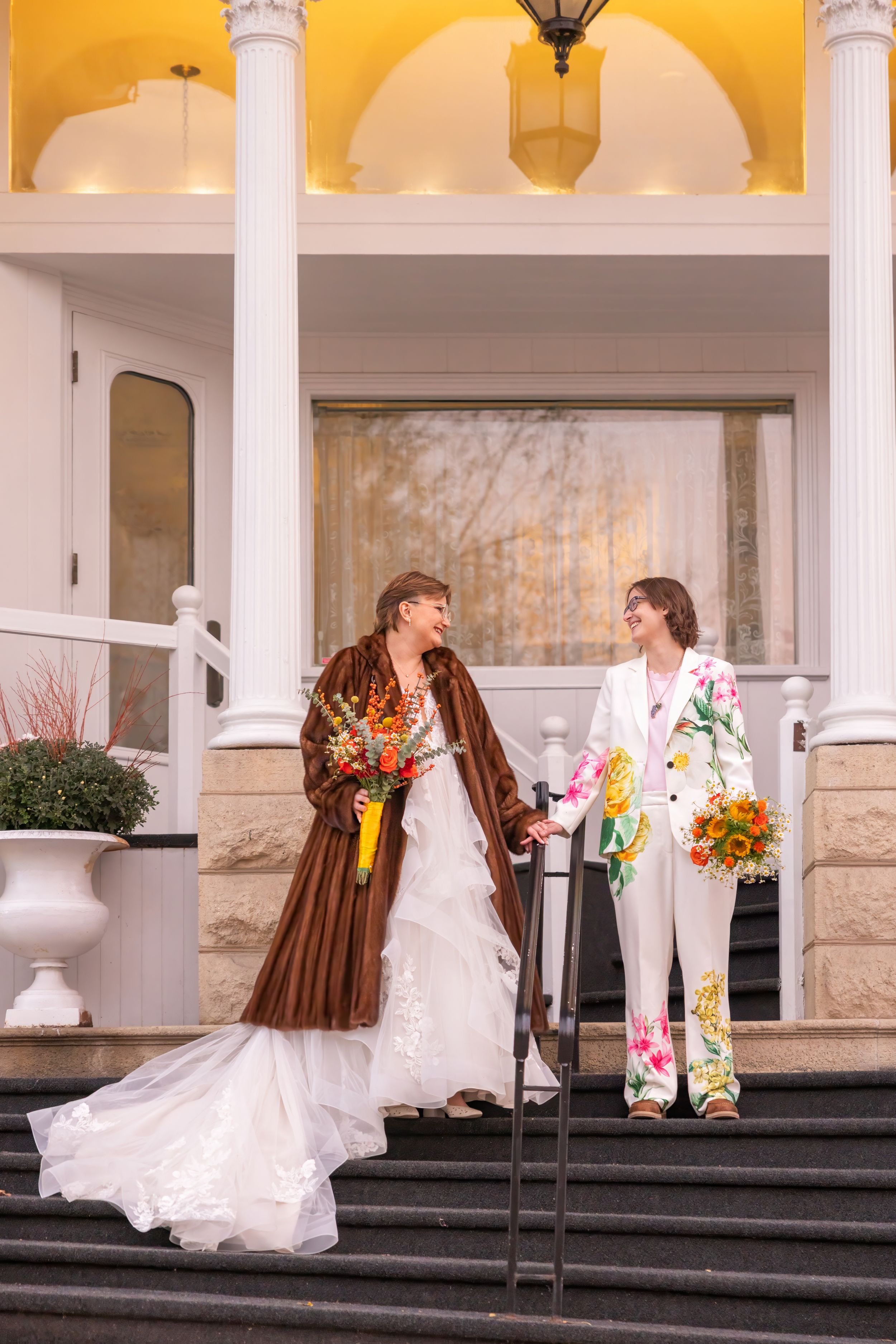 A wedding couple poses on stone steps outside a white columned building with yellow walls.