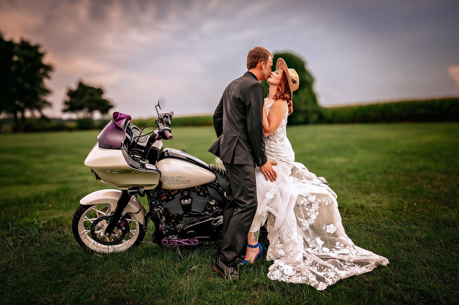 A bride in a white lace wedding dress with a red bouquet poses with her groom along a brick building exterior.