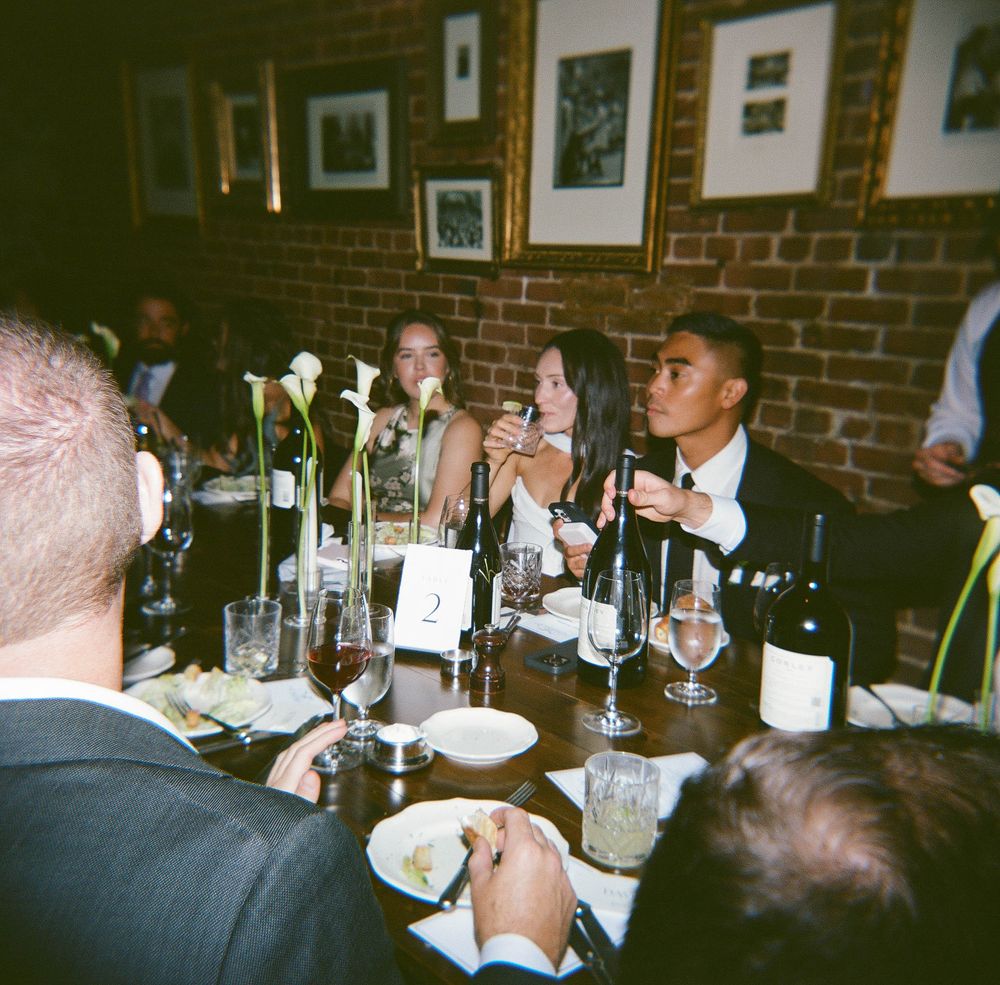 People gathered around a table with wine glasses and bottles in a dimly lit restaurant with brick walls and framed photos.