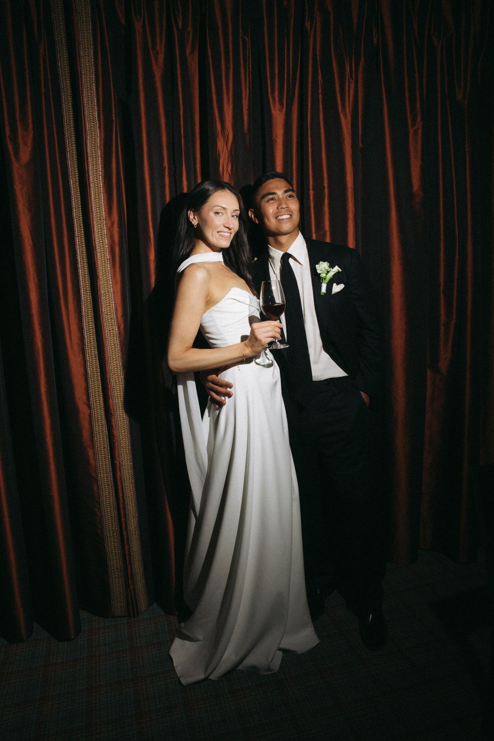A couple in formal wedding attire share an intimate moment against a dark red curtain backdrop.