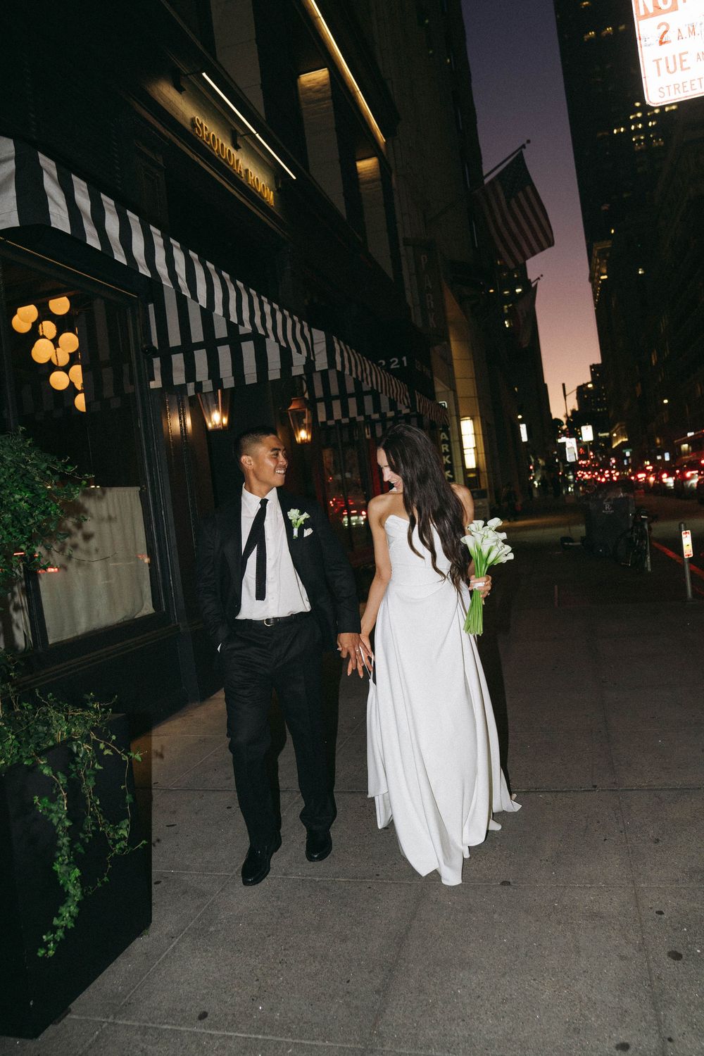 A couple in wedding attire walks down a city sidewalk at dusk with illuminated storefronts in the background.