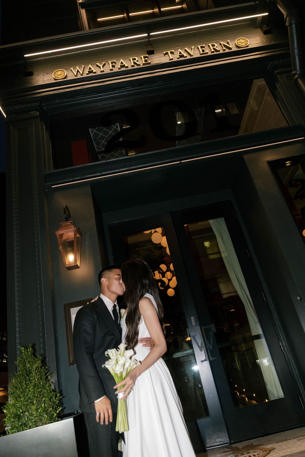 A romantic wedding couple shares an intimate moment outside a venue entrance with dramatic lighting and signage.