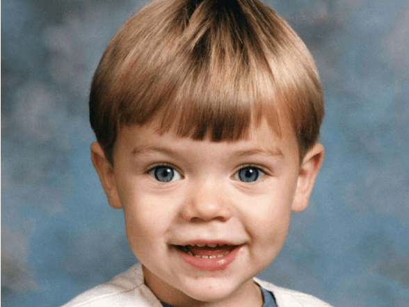 A young child with brown hair and bright eyes smiles warmly against a mottled blue studio backdrop.