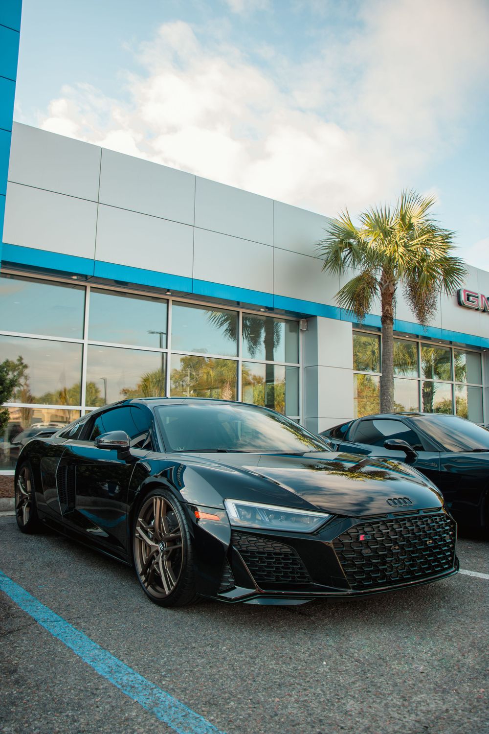 Black Audi R8 sports car parked in front of a modern dealership with palm trees and blue accents.