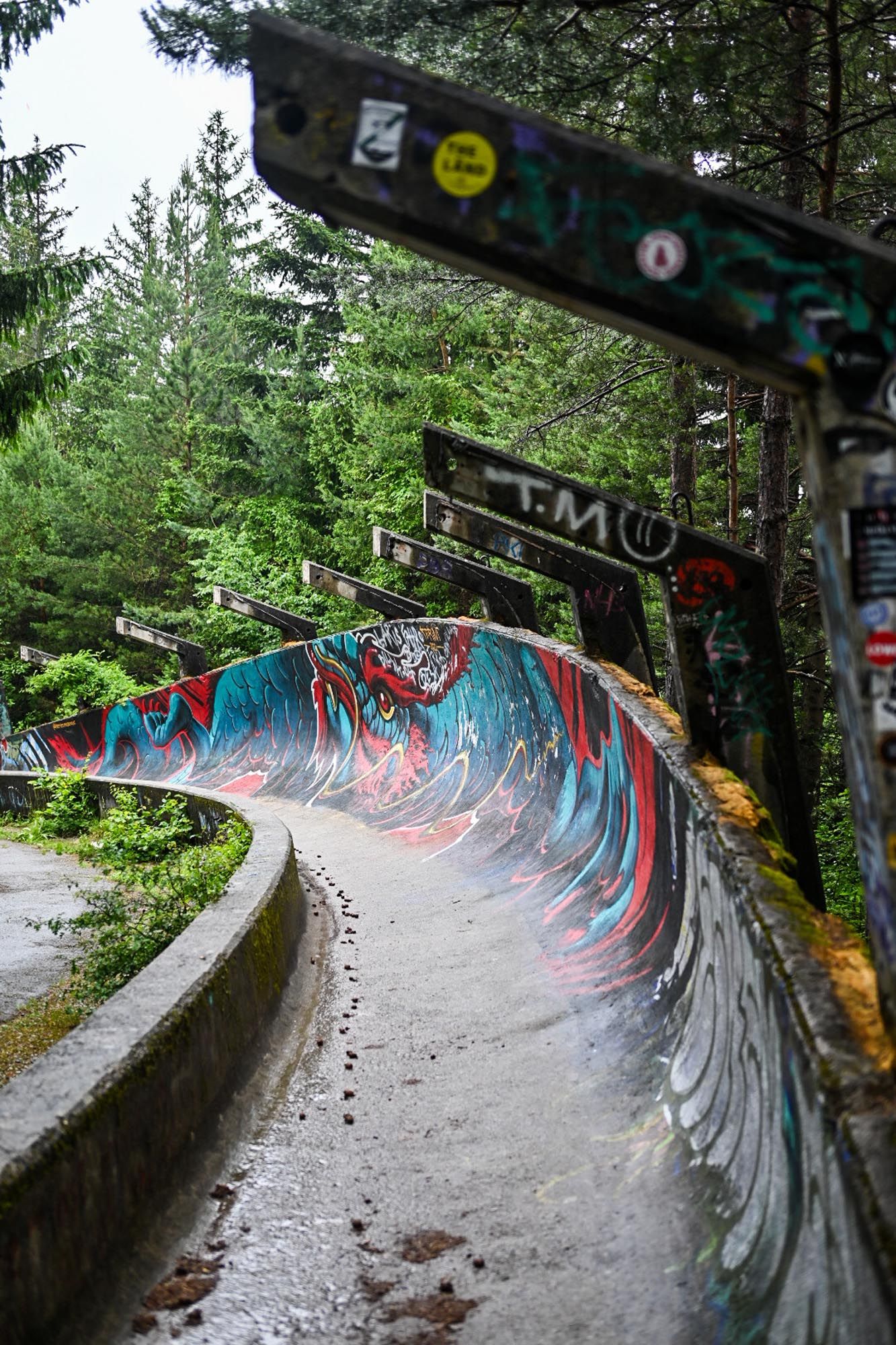 Graffiti-covered abandoned concrete bobsled track curves through forest with colorful street art on its weathered walls.