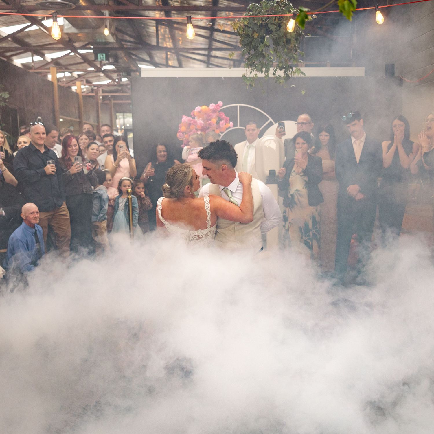 A crowd gathers at an indoor venue as dramatic fog or smoke effects fill the space during the bride and groom first dance.