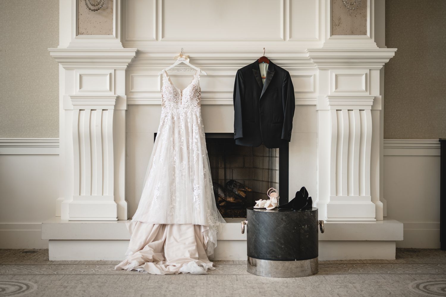 Wedding dress and tuxedo displayed on hangers against an elegant white fireplace mantel.