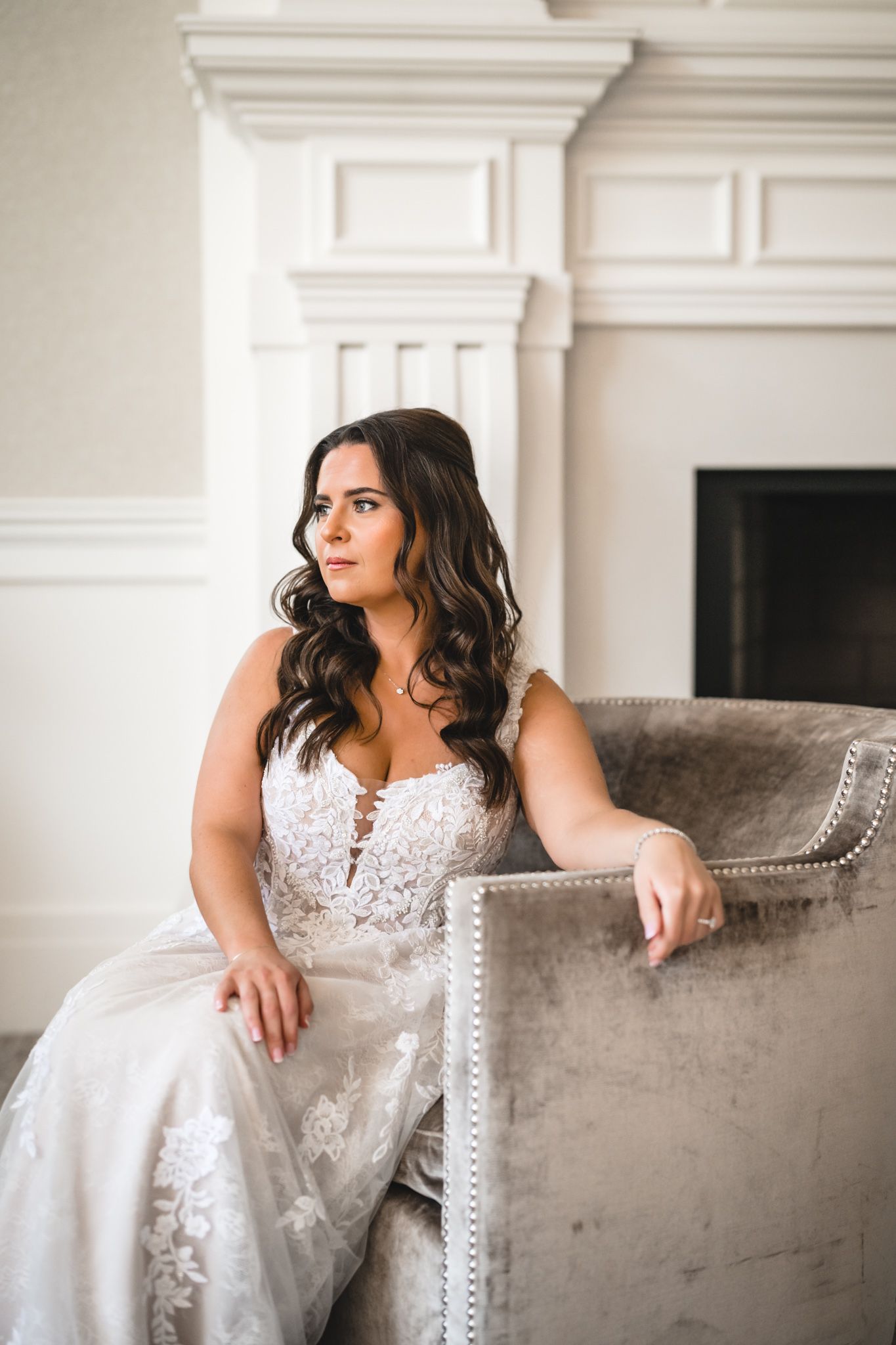 A bride in a lace wedding gown sits elegantly on a velvet chair near a white fireplace.