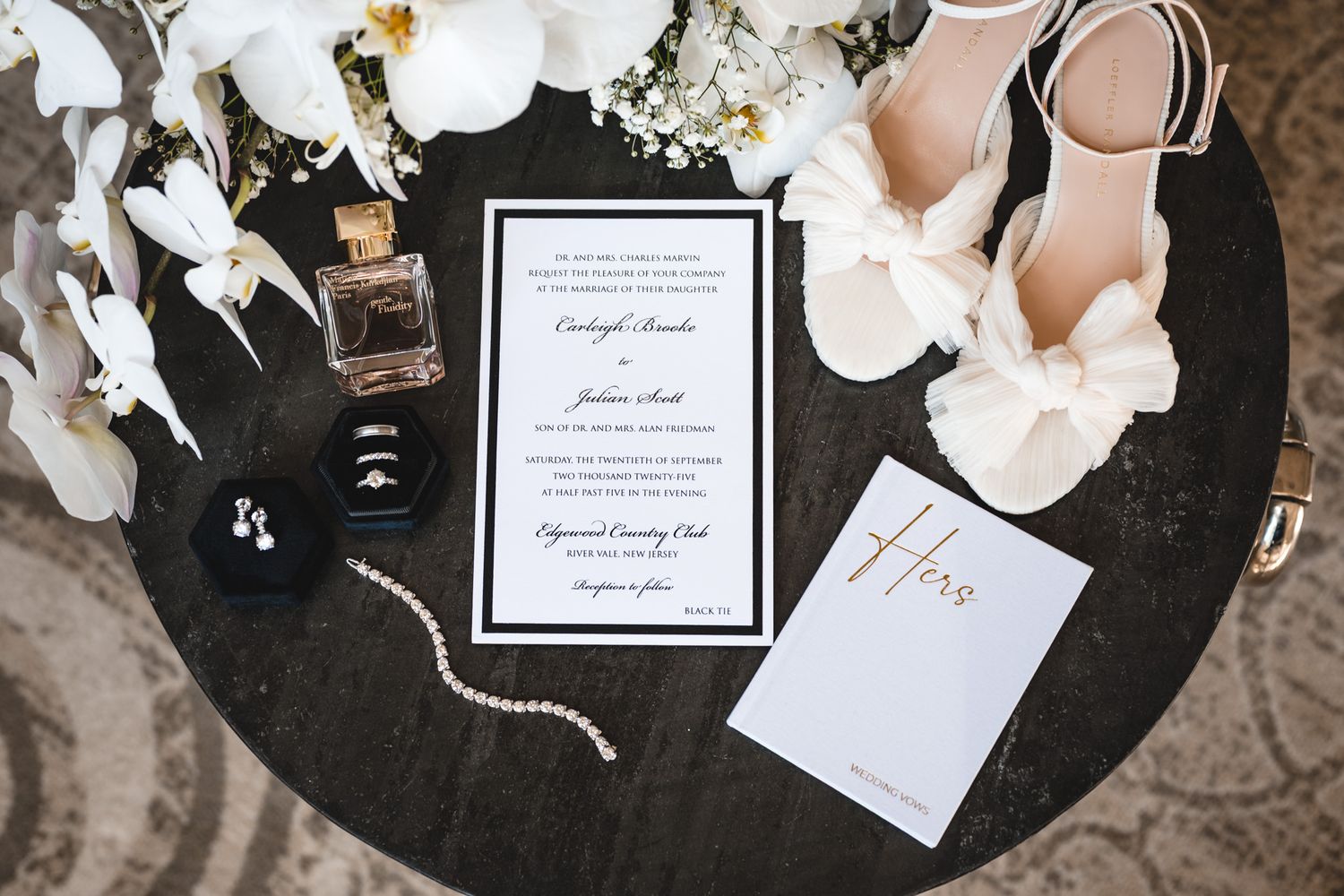 Wedding invitation suite with white orchids, perfume, and accessories on dark table.