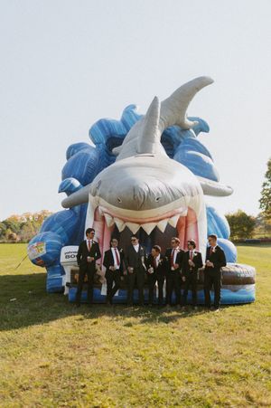 Wedding party poses in front of a large inflatable shark decoration on a grassy field.