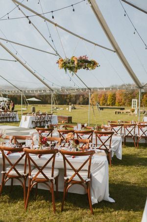 Elegant wedding reception setup under a white tent with wooden cross-back chairs and string lights.