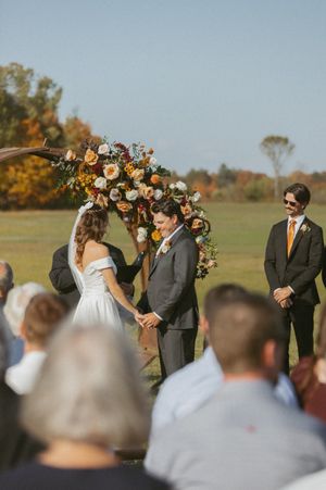 An outdoor autumn wedding ceremony with fall foliage and floral arch decorations.