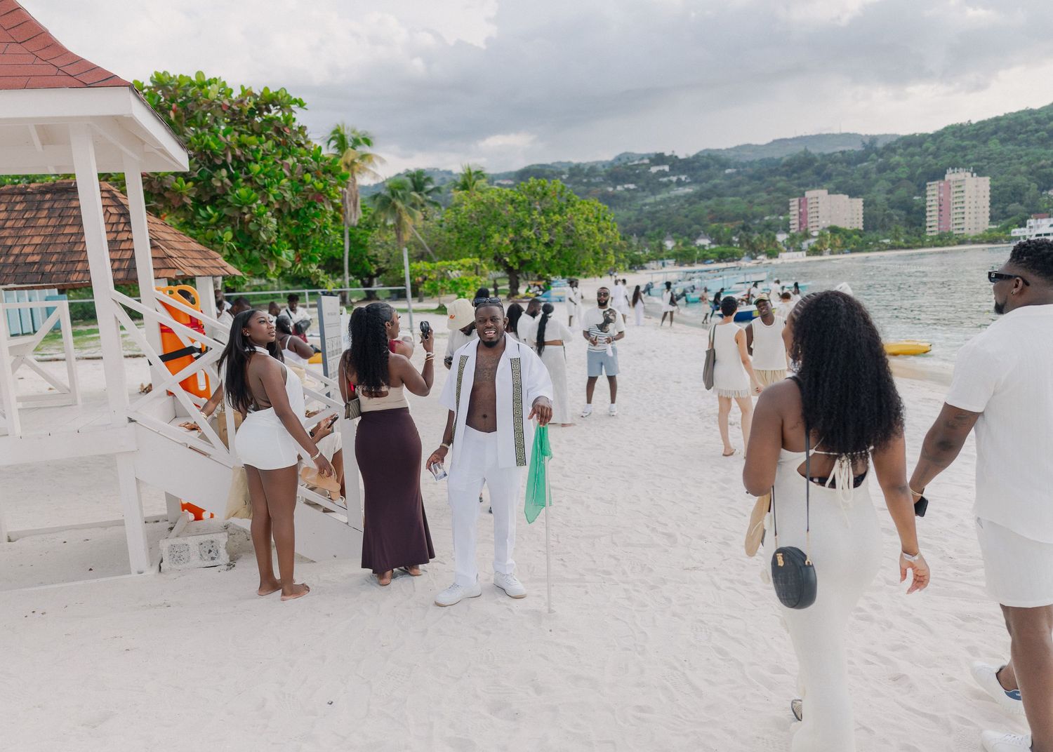People gathered on a scenic white sandy beach with mountains and buildings visible in the background.