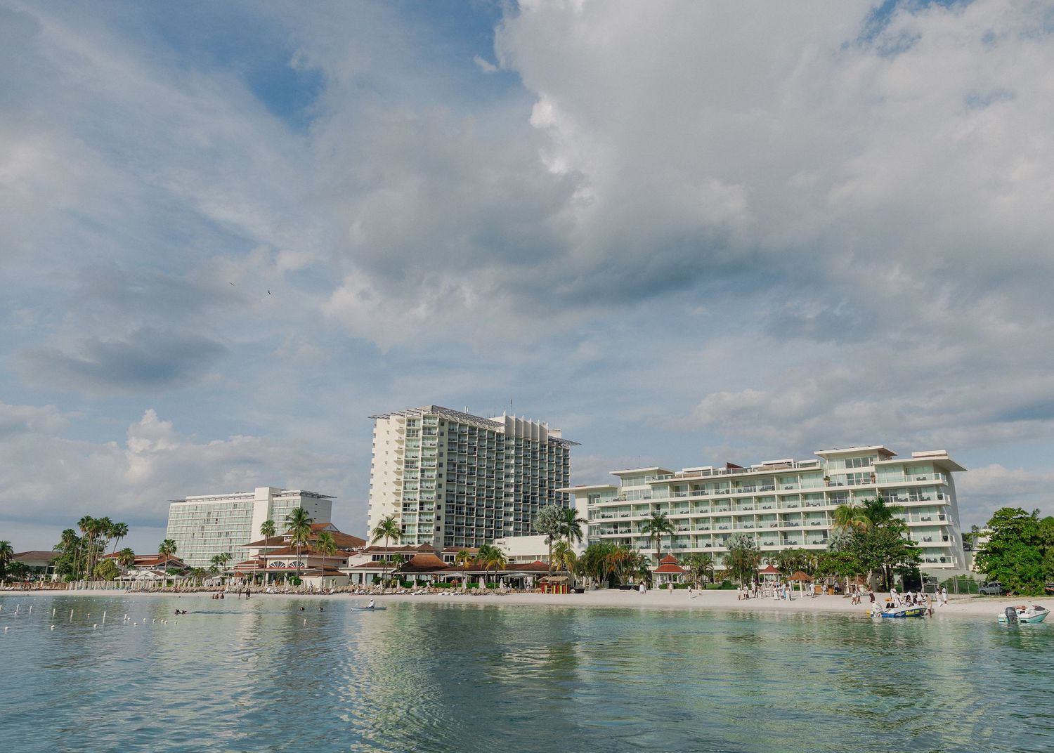 Panoramic view of a beachfront resort complex with hotels and palm trees along the shoreline under cloudy skies.