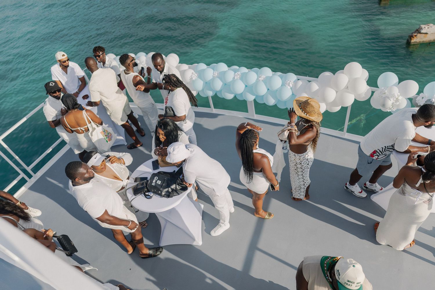 A group of people in white outfits sit on steps near turquoise water during a seaside celebration.