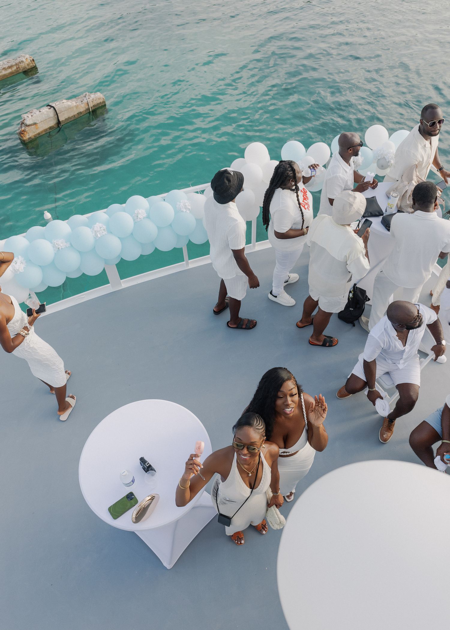 People in white attire gather on a boat deck surrounded by white balloons against turquoise waters.