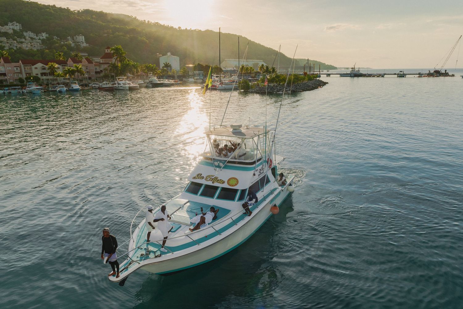 Aerial view of a fishing boat cruising in calm waters at sunset with mountains and harbor in background.