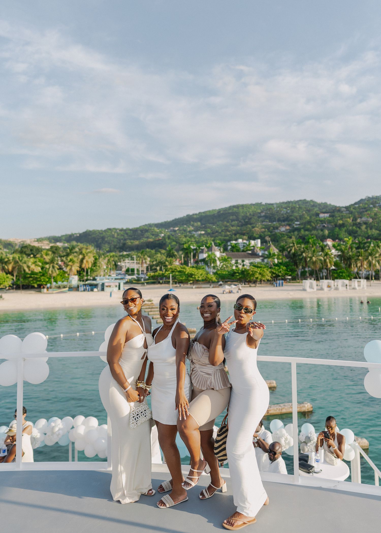 Group in white outfits posing together on a boat deck with tropical beach and palm trees in the background.