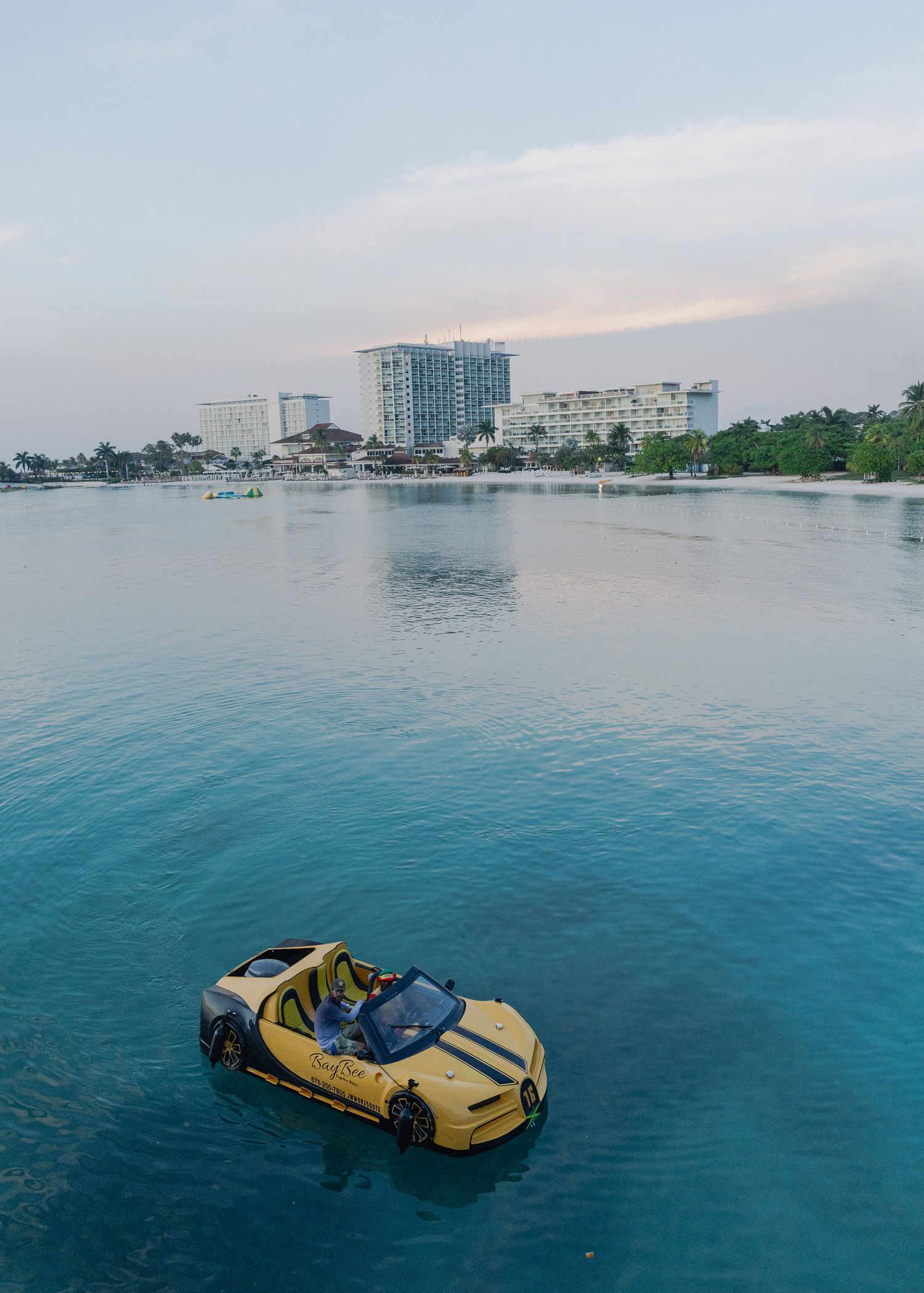 A yellow sports car appears to be floating or submerged in blue ocean waters with coastal buildings visible in the background.