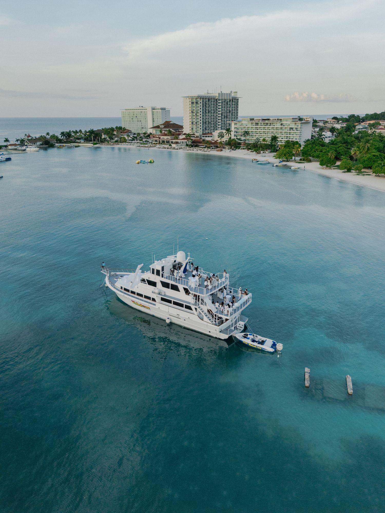 Aerial view of a large yacht cruising in turquoise waters near a coastal resort with high-rise buildings.