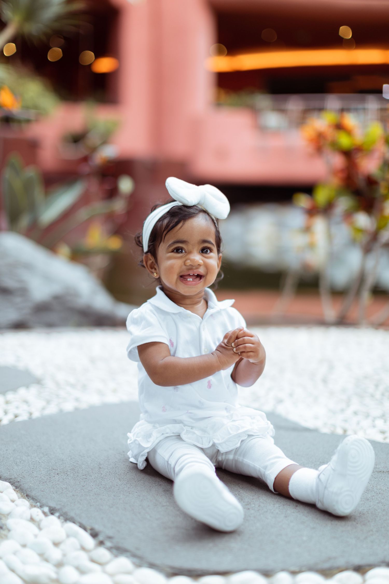 Small child in all-white outfit with bow sitting on gray surface.