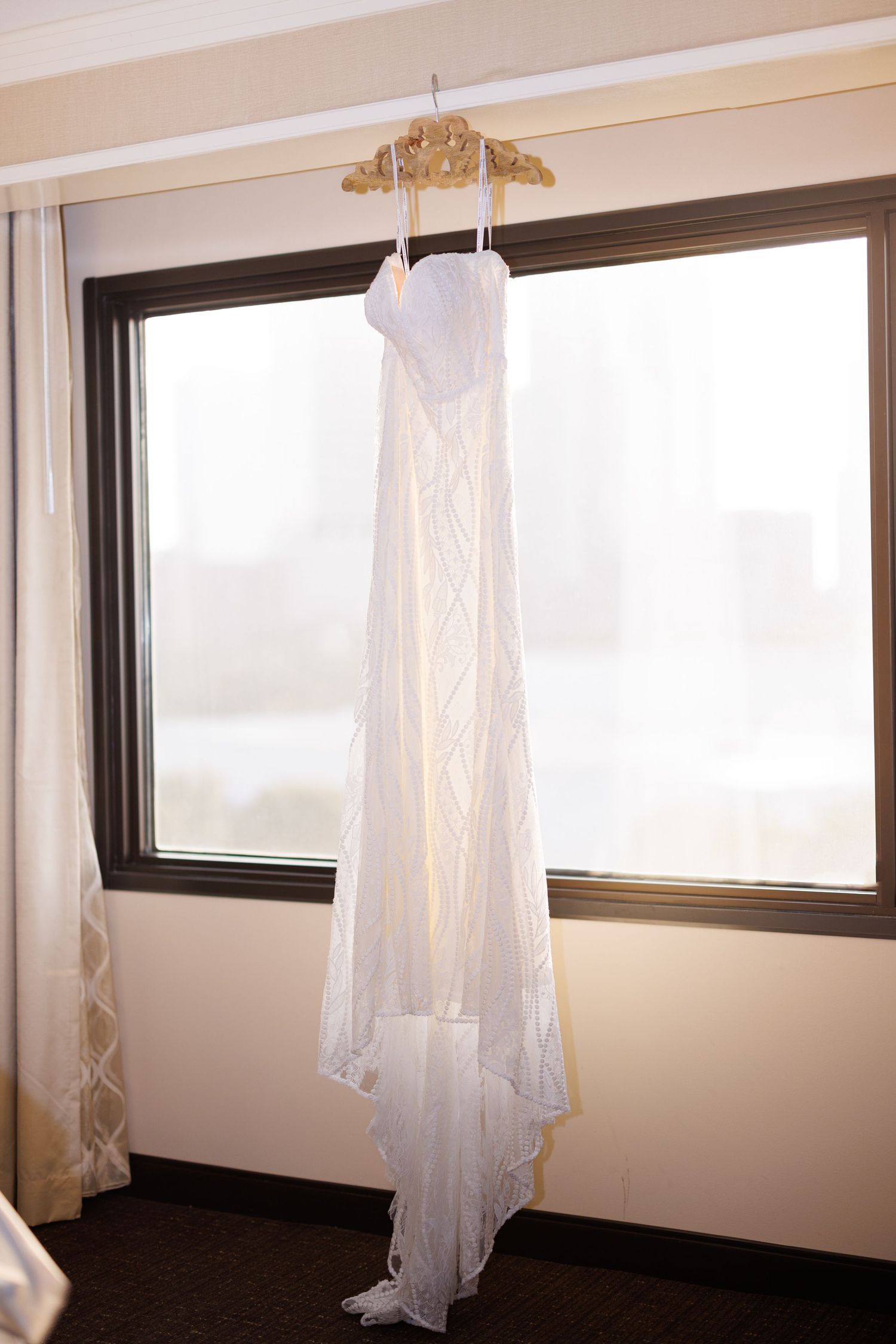 A white wedding dress hangs in a hotel room window with natural lighting.
