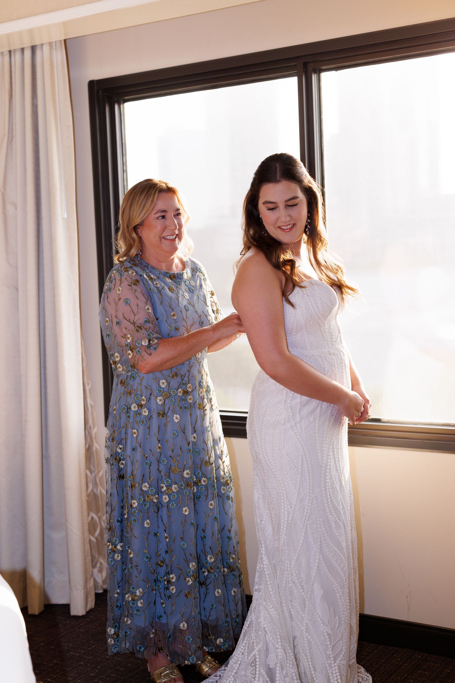 Two people in floral and white dresses share a joyful moment in a hotel room.