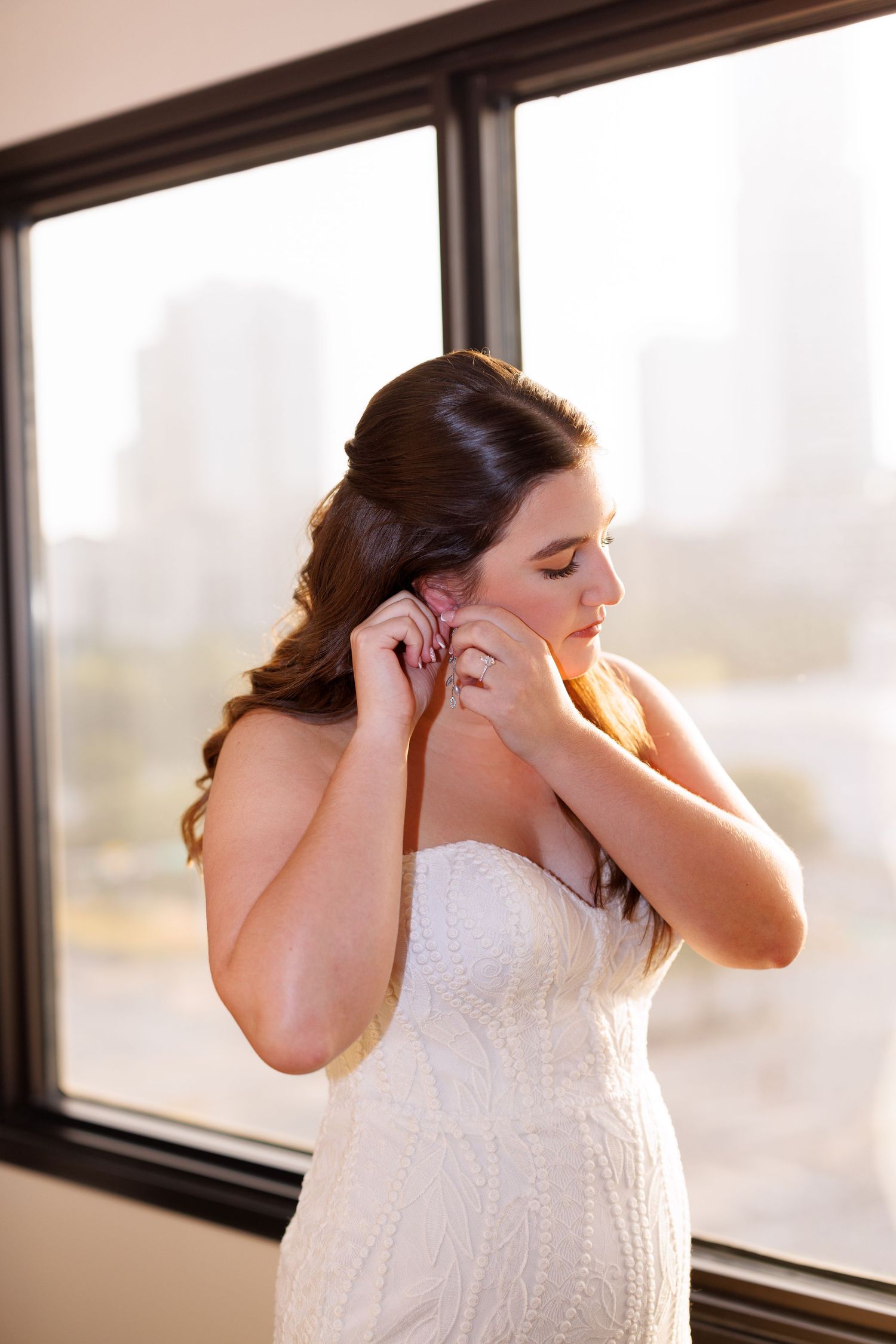 A bride in a white lace wedding dress adjusts her earrings while standing by a window in natural light.