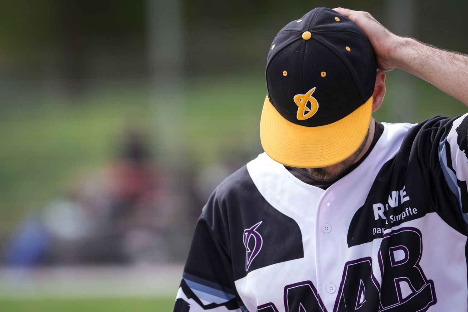 Baseball player in black and yellow cap with DAAB jersey scratching head on field.