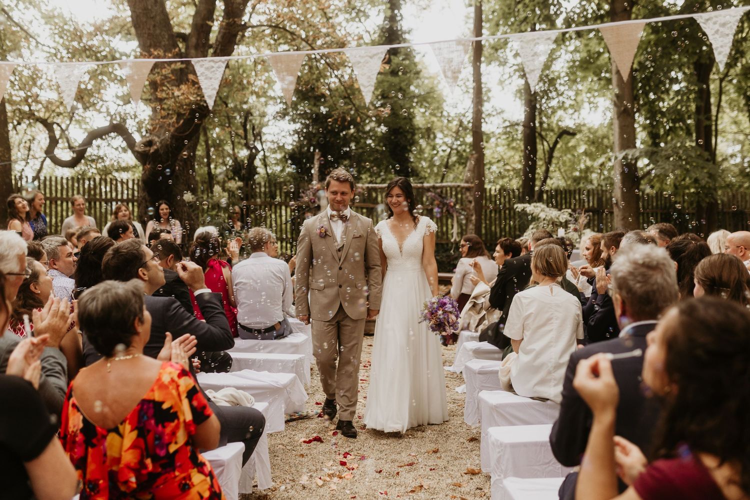 Bride and groom raise hands in celebration at outdoor woodland wedding ceremony with bunting and petal-lined aisle.