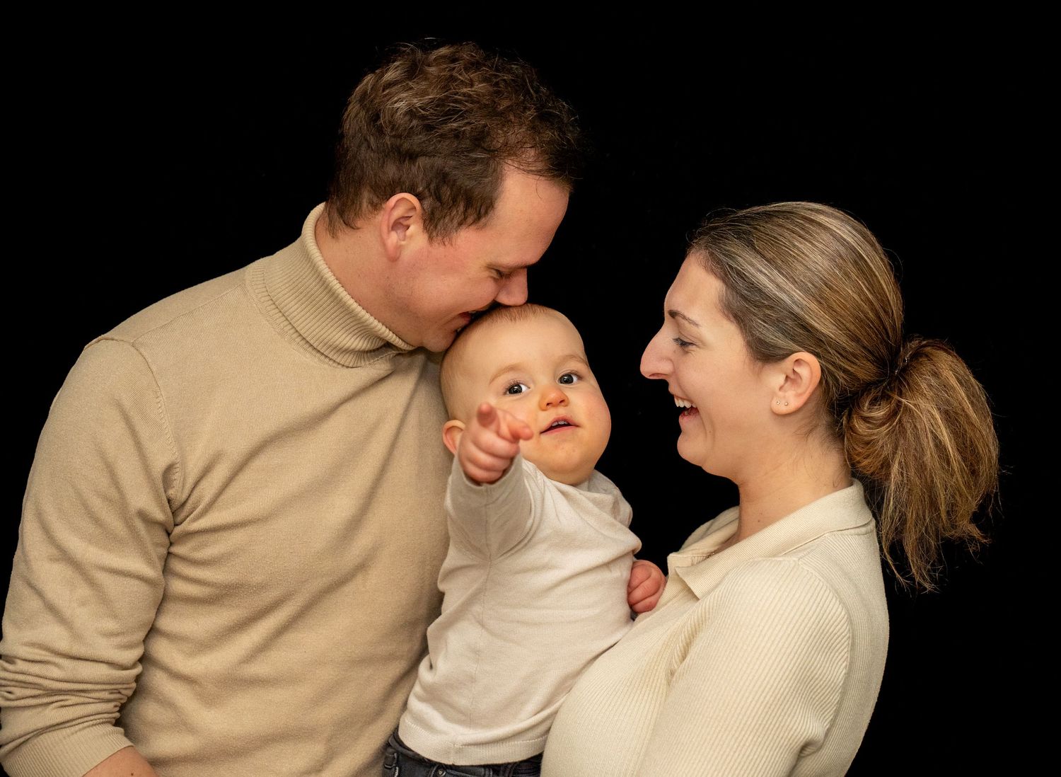 Ouders in crèmekleurige truien kussen en glimlachen naar een baby met een geschminkt gezichtje op een zwarte achtergrond.