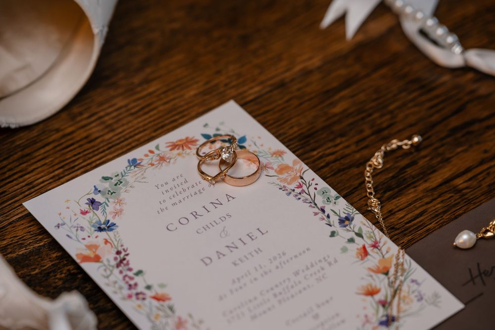 Wedding invitation for Corina and Daniel adorned with floral border, gold rings, and jewelry on wooden table.