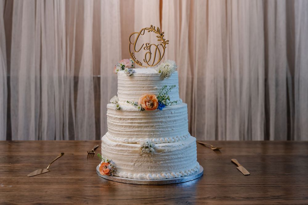 Three-tier white wedding cake with floral accents and gold monogram topper on wooden table.