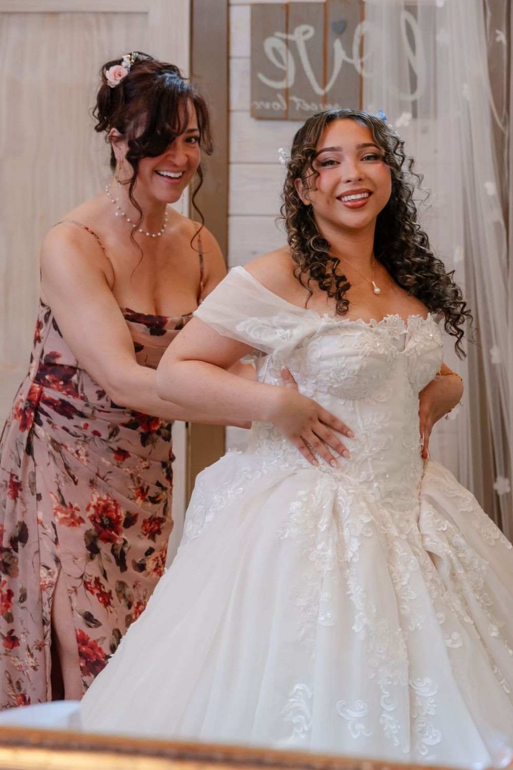 A smiling woman in a floral dress helps a bride in an off-shoulder white ball gown at a bridal boutique.