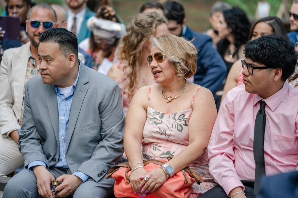 Three wedding guests seated outdoors, smiling and enjoying a ceremony on a sunny day.
