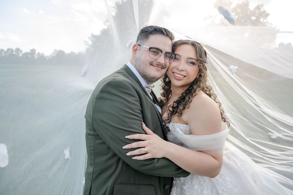 Bride and groom embracing under flowing white veil outdoors on wedding day.