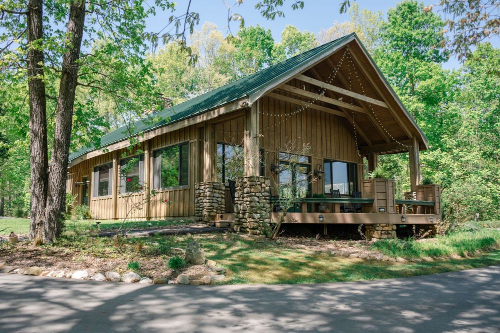 Rustic wooden cabin with green roof surrounded by lush trees, featuring a covered porch with stone accents and large windows.