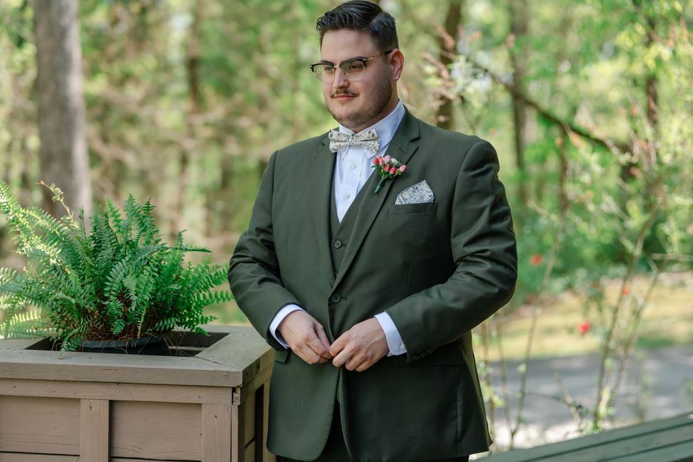 Groom in olive green suit with floral boutonniere adjusting jacket outdoors near ferns.