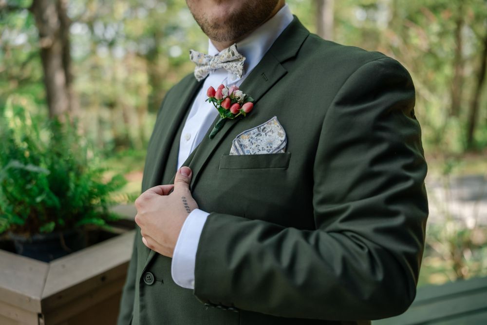 Groom in olive green suit with floral boutonniere and paisley pocket square adjusting jacket outdoors.