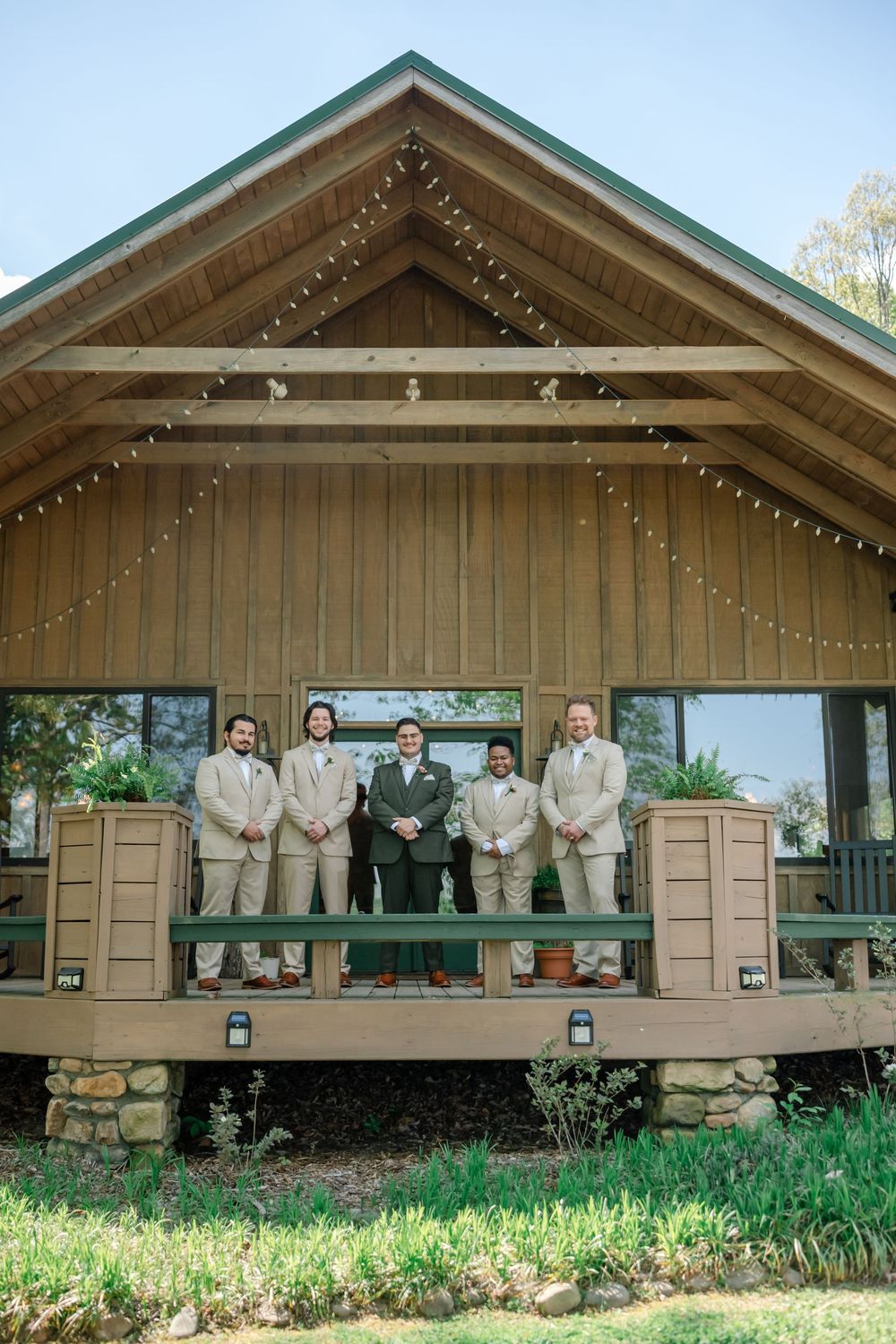 Groom and four groomsmen in tan suits posing on a wooden cabin porch decorated with string lights.