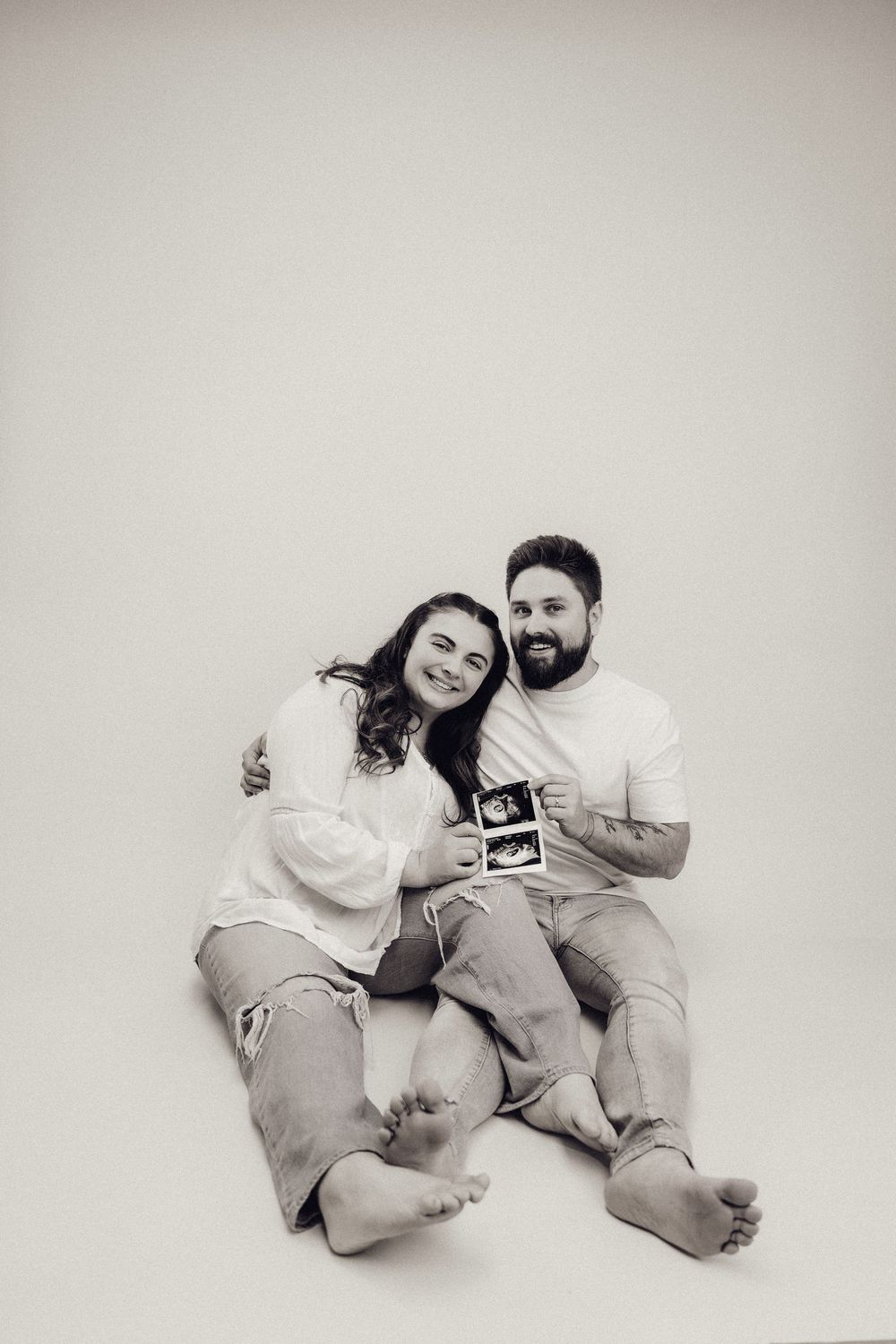 Black and white photo of smiling couple sitting on floor holding ultrasound images.