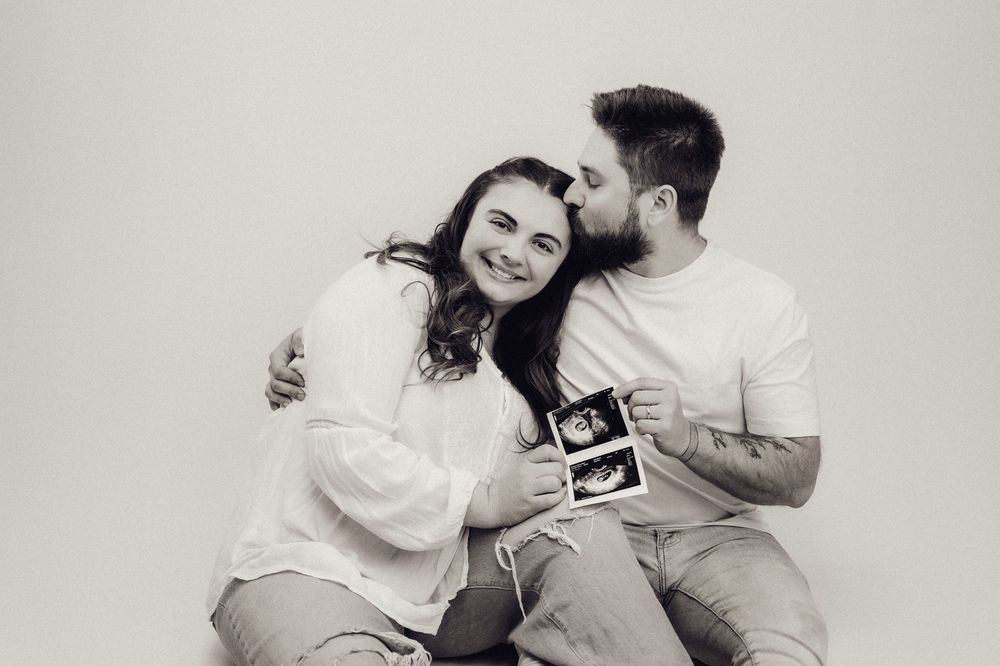 Black and white photo of a happy couple sitting together holding ultrasound scans, man kissing woman on cheek.