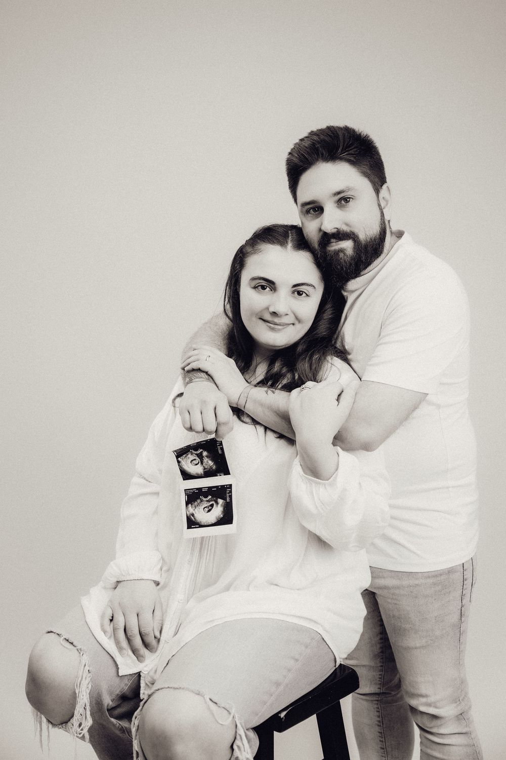 Black and white maternity photo of a couple, man standing behind seated woman holding ultrasound images.