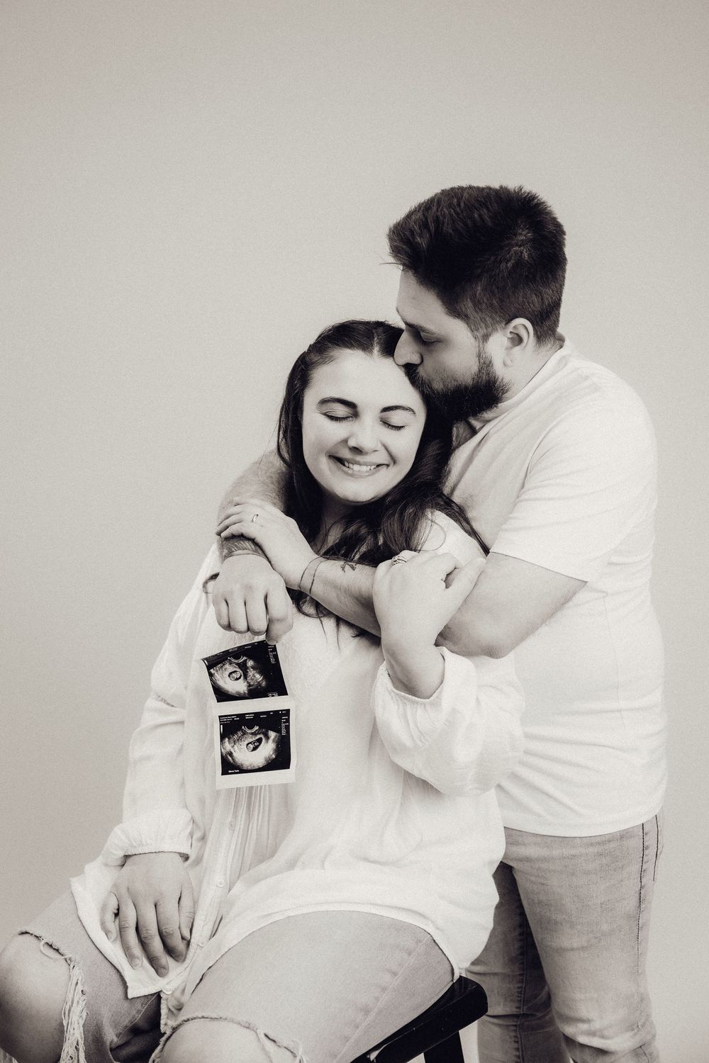 Black and white photo of a bearded man kissing smiling pregnant woman on head while she holds ultrasound scans.